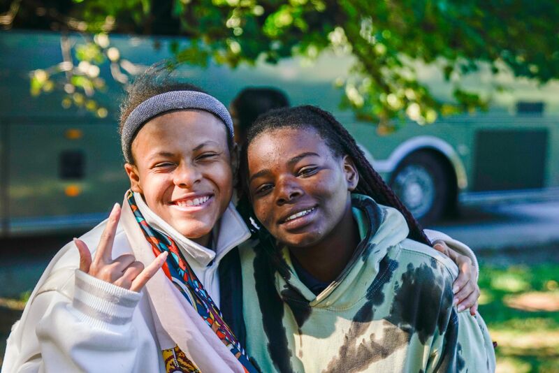 The image shows two young women posing closely together, likely friends. The woman on the left is wearing a gray headband and a patterned scarf, flashing a hand gesture. The woman on the right has dreadlocks and is wearing a green and black hoodie. They both have warm smiles and appear to be outdoors, with a vehicle visible in the background.